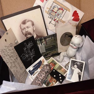 Könnte beinhalten: Eine Schachtel gefüllt mit Vintage-Ephemera, darunter Fotos, Postkarten, ein handgeschriebener Brief, ein Domino und eine kleine Porzellanpuppe. Die Schachtel ist mit weißem Seidenpapier ausgekleidet und hat einen roten Samt-Boden.