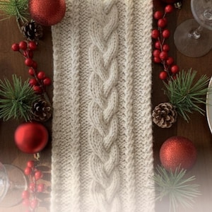 May include: A cream-colored knit cable braid table runner centered on a wooden surface, flanked by white plates, clear wine glasses, red ornaments, pine sprigs, and small pinecones. The text "Knit Cable Braid Table Runner" is visible.