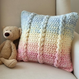 May include: A handmade, gradient-colored crochet pillow in shades of pink, yellow, and blue, with a cable knit design. A plush teddy bear sits next to the pillow on a cream-colored armchair. The pillow is a decorative item.