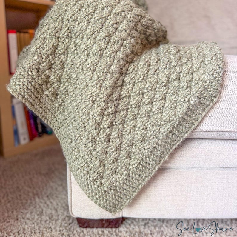 May include: A sage green hand-crocheted blanket featuring a thick, textured cable knit pattern. The yarn is a soft, muted olive tone with a consistent stitch throughout the fabric, creating a dense and warm appearance for this textile item.