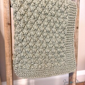 May include: A sage green knitted blanket featuring a textured diamond pattern across the centre and a garter stitch border. The fabric has a thick, soft appearance with visible yarn loops. The text See Love Share is printed in the bottom right corner.
