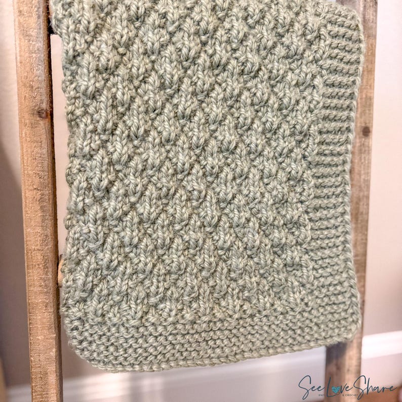 May include: A sage green knitted blanket featuring a textured diamond pattern across the centre and a garter stitch border. The fabric has a thick, soft appearance with visible yarn loops. The text See Love Share is printed in the bottom right corner.