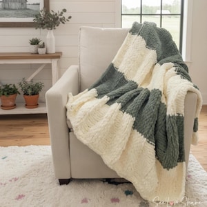 Puede incluir: Una manta de punto grueso, con rayas crema y verde apagado, sobre un sillón crema. La textura y la combinación de colores de la manta crean una sensación acogedora. Un accesorio decorativo para el hogar.