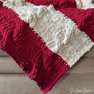 May include: A cozy, hand-knitted blanket in a cable-knit design. The blanket features alternating sections of vibrant red and creamy white, creating a striking visual contrast. The thick yarn gives it a plush, inviting texture.
