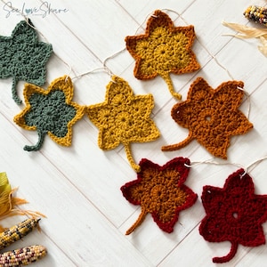 Puede incluir: Una guirnalda de hojas de otoño tejidas a crochet en tonos de verde, amarillo, naranja y rojo. Las hojas están enhebradas con un cordel y tienen un diseño simple y rústico.