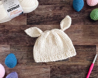 Könnte beinhalten: Eine weiße gestrickte Baby-Mütze mit Hasenohren. Die Mütze ist aus Bernat Softee Chunky Garn gefertigt. Die Mütze ist von bunten gehäkelten Ostereiern umgeben.