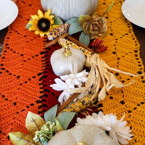 May include: A crocheted table runner in orange, red, and yellow with a white, gray, and gold pumpkin, corn husks, sunflowers, and white flowers.