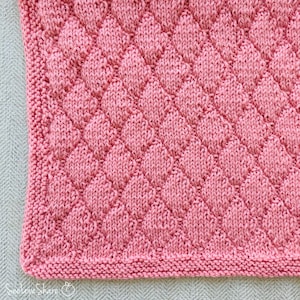 May include: Close-up of a pink knitted blanket with a textured diamond pattern. The blanket has a ribbed border and is made of soft yarn. The diamond pattern creates a raised, dimensional effect. The blanket is a cozy, handmade item.