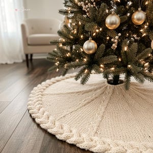 Könnte beinhalten: Ein kreisförmiger, cremefarbener gestrickter Weihnachtsbaumteppich mit einem dicken Zopfmuster-Rand und einer gerippten Textur, die von der Mitte ausstrahlt. Der Stoff besteht aus grobem Strickgarn und bildet eine weiche, strukturierte Basis.