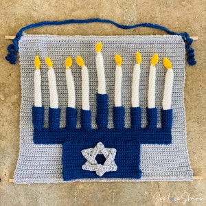 Puede incluir: Un tapiz de pared de menorá tejido a crochet con hilo azul y gris. La menorá tiene nueve velas, cada una con una llama amarilla. Una estrella de David plateada está en el centro de la menorá.