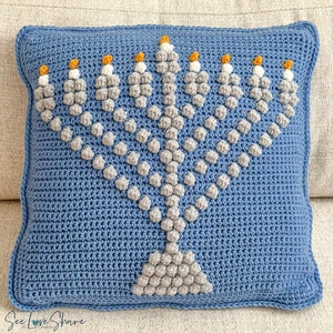 May include: A square, blue crochet cushion featuring a grey menorah design with yellow accents. The menorah is made with textured stitches, and the cushion has a simple border. The text "See Love Share" is visible in the bottom left corner.