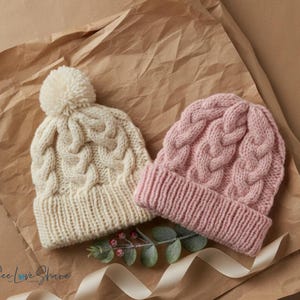 Peut inclure: Deux bonnets en tricot torsadé, un crème avec un pompon, l'autre rose. Les deux ont un bord côtelé et un motif torsadé. Ils reposent sur du papier brun avec de la verdure et un ruban.