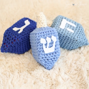May include: Three crocheted dreidels, one blue with a white Hebrew letter "Shin", one blue with a white Hebrew letter "Nun", and one blue with a white Hebrew letter "Gimel", on a light brown fuzzy surface.