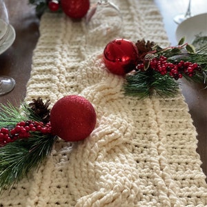 Puede incluir: Un camino de mesa de crochet blanco con un patrón de punto de cable está decorado con adornos rojos y ramitas de vegetación con bayas rojas.