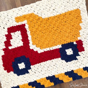 May include: A crocheted blanket depicting a red truck with a yellow load bed and blue wheels. The truck is on a white background, with a border of blue and yellow stripes. The blanket is made with soft yarn, suitable for a child's room.