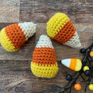 May include: Three crocheted candy corn decorations featuring a triangular shape with horizontal bands of yellow, orange, and white yarn. A small foam candy corn piece sits near a black decorative branch with small yellow and orange berries.