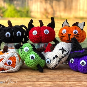 May include: A collection of seven crocheted Halloween-themed pumpkins, each with googly eyes and unique features. The pumpkins are in various colors, including black, red, orange, green, white, and purple.  SeeLoveShare is printed on the wooden surface.