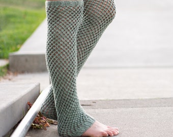 May include: A pair of sage green knitted leg warmers with a fishnet pattern. The leg warmers are worn on a person's legs.