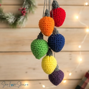 May include: A hanging decoration featuring six crocheted light bulb shapes in red, orange, green, blue, yellow, and purple. Each bulb has a grey crocheted base and is suspended by thin grey strings. The text See Love Share is visible at the bottom.