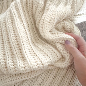 May include: Cream-coloured crochet blanket with a ribbed pattern. The blanket is folded, showing the texture and stitch detail. The text "Beginner Crochet Pattern" is visible.