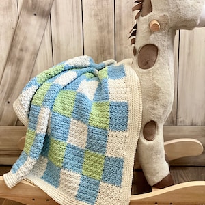 May include: A crocheted baby blanket with a checkered pattern in shades of blue, green, and white. The blanket is draped over a rocking horse.