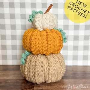 Op de afbeelding: Een stapel van drie gehaakte pompoenen in beige, oranje en wit met groene gehaakte bladeren. De pompoenen zijn op elkaar gestapeld en staan op een houten oppervlak. De tekst "NEW CROCHET PATTERN" staat in een gele cirkel in de rechterbovenhoek van de afbeelding.