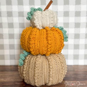 May include: A stack of three crocheted pumpkins in beige, orange, and white with green leaves. The pumpkins are stacked on top of each other, with the largest pumpkin on the bottom and the smallest pumpkin on top. The pumpkins are decorated with green leaves and a cinnamon stick.