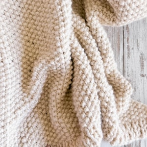 May include: A chunky, off-white hand-knitted blanket featuring a consistent garter stitch texture. The fabric is thick and soft with visible yarn loops. In the bottom right corner, the text See Love Share is printed in a dark blue script font.