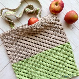 May include: A handmade crochet tote bag in beige and light green, with matching handles. The bag is filled with red apples. The open weave design is visible. The bag is designed for carrying groceries or other items.
