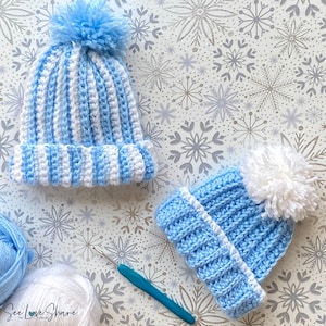May include: Two crocheted hats with pom-poms in blue and white stripes. The hats are on a white background with a snowflake pattern. A crochet hook is also in the image.