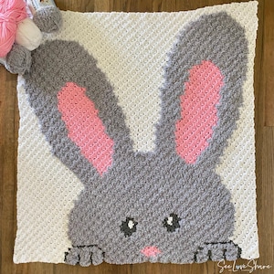 Puede incluir: Una manta de bebé de crochet con una cara de conejo gris en el centro. El conejo tiene orejas rosas y una nariz rosa. La manta es blanca con un borde gris.