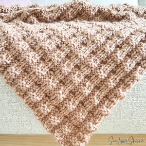 May include: A close-up of a brown knitted blanket with a textured pattern. The blanket is folded and draped over a light-colored surface.