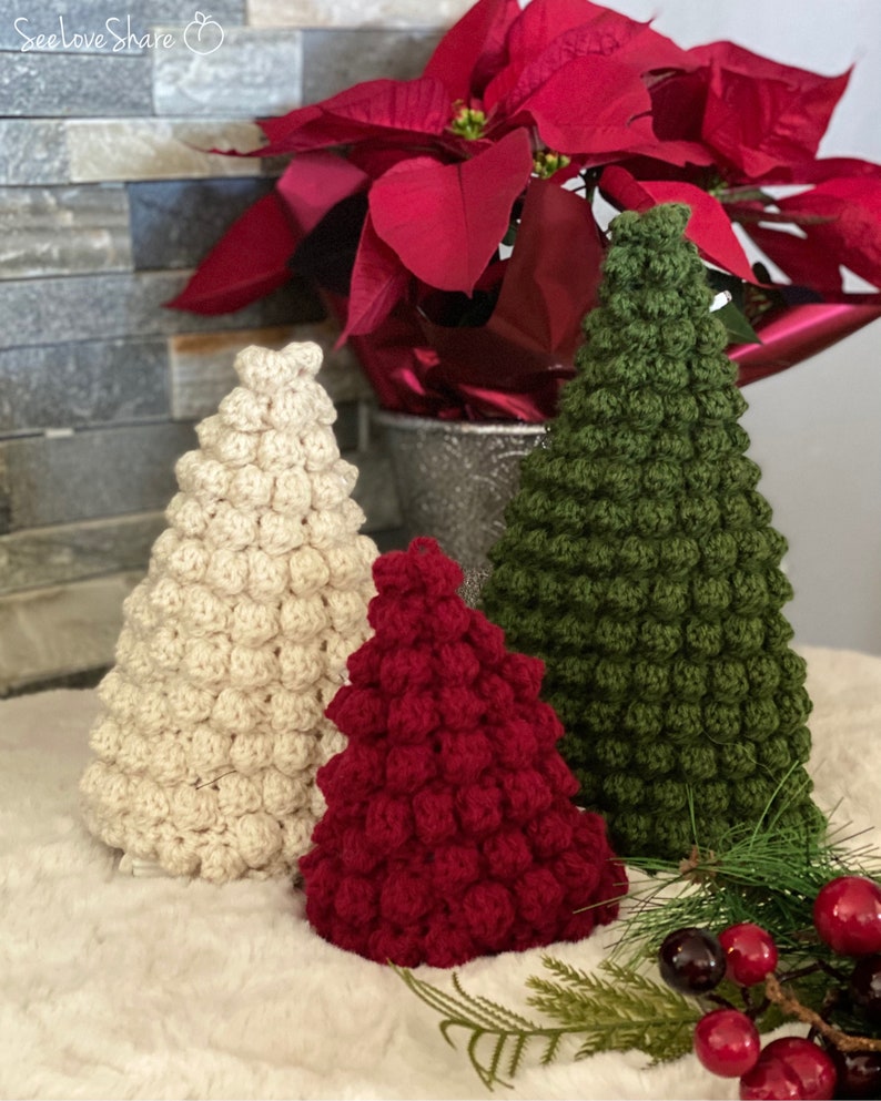 May include: Three crocheted Christmas trees in cream, dark red, and dark green.  The trees are cone-shaped and feature a textured, bobble stitch. They are arranged on a white faux fur surface in front of a poinsettia plant.  SeeLoveShare logo is visible in the upper left corner.