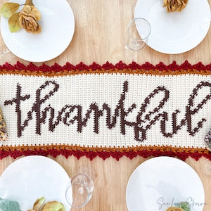 May include: A cream-colored crochet table runner with the word "thankful" in brown script. The runner has a scalloped, dark red border and is set on a wooden table with white plates, glasses, and dried flowers, creating a fall-themed tablescape.