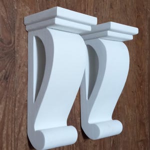 Puede incluir: Dos ménsulas de madera blancas con un diseño decorativo de volutas. Las ménsulas tienen una parte superior en capas y una base curva y de soporte. Están montadas sobre una superficie de madera marrón, mostrando sus detalles arquitectónicos.