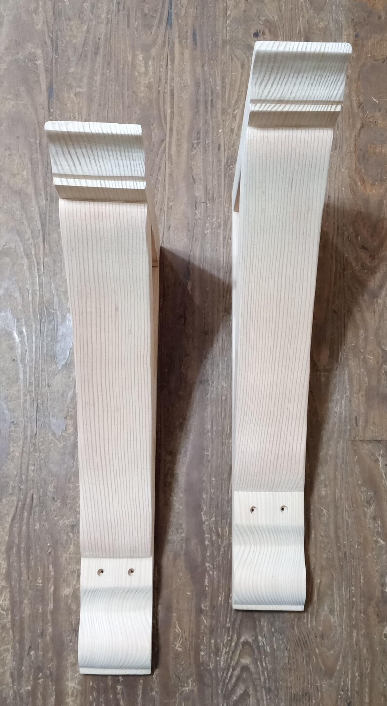 May include: Two unfinished wooden corbels with a light, natural wood grain. Each corbel has a decorative top and bottom, with a rectangular body. The corbels are designed for structural support and add a decorative element.