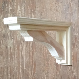 Puede incluir: Un soporte de estante de madera sin terminar, listo para pintar o teñir. Cuenta con un estante rectangular sostenido por un soporte curvo y una pieza de montaje vertical. La veta de la madera es visible, mostrando el material natural.