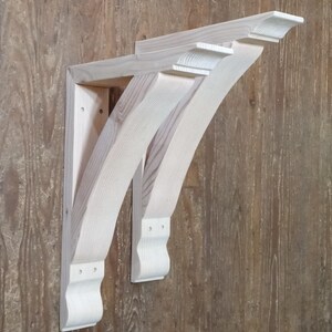 May include: Two unfinished wooden corbels, with a curved design and decorative detailing. The corbels are light in colour and appear to be made of pine or similar wood. They are mounted on a wooden surface, showcasing their architectural style.