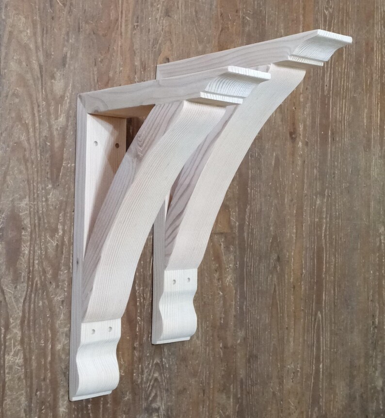 May include: Two unfinished wooden corbels, with a curved design and decorative detailing. The corbels are light in colour and appear to be made of pine or similar wood. They are mounted on a wooden surface, showcasing their architectural style.