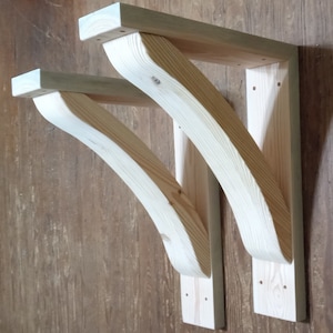 May include: Two unfinished wooden shelf brackets. Each bracket has a rectangular top piece, a curved support, and a vertical mounting piece. The wood grain is visible, and the brackets are ready for staining or painting.