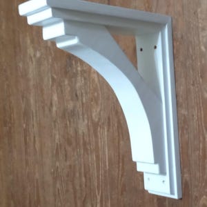 Puede incluir: Un soporte de estante de madera blanco con un diseño escalonado. El soporte tiene un soporte arqueado y está montado sobre una superficie de madera. El soporte está diseñado para soportar un estante.