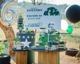 May include: A military-themed birthday party setup featuring a large sign reading TROOPS ATTENTION WELCOME TO BASECAMP IT'S RUBEN'S 6TH BIRTHDAY, a silver number 6 balloon, green and black balloons, cakes, cupcakes, and wooden spool tables.