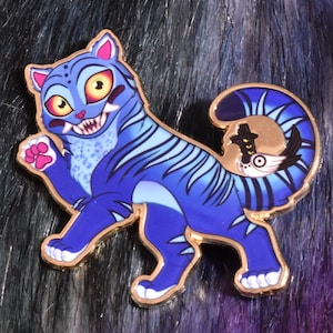 以下が含まれることがあります： 金色の縁取りが付いた青い猫の形をしたエナメルピン。猫は黄色い目と歯を見せた笑顔で、恐ろしい表情をしています。ピンには青い縞模様と尾にデザインが施されています。背景は黒と紫のフェイクファーのミックスです。
