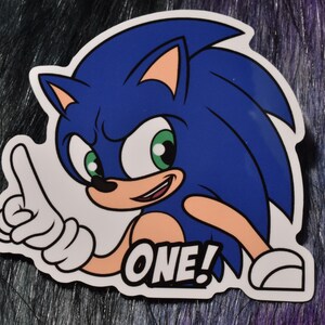 May include: A sticker featuring Sonic the Hedgehog, a blue anthropomorphic hedgehog. Sonic is pointing with one finger and smiling. The word "ONE!" is printed below him in a white box with a black outline. The sticker has a white border.