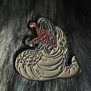 May include: A detailed enamel pin featuring a stylized creature design. The pin has a light gray body with a dark gray outline and a red and dark gray creature in the center. The pin is set against a dark, textured background.