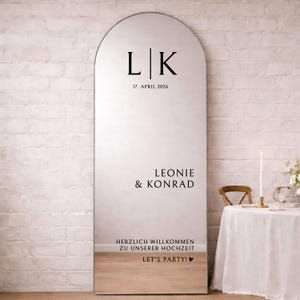 Könnte beinhalten: Ein hoher, gewölbter Standspiegel mit schwarzer, individueller Beschriftung. Das Design zeigt die Initialen L|K, das Datum 17. APRIL 2026, die Namen LEONIE & KONRAD sowie den Text HERZLICH WILLKOMMEN ZU UNSERER HOCHZEIT und LET'S PARTY!