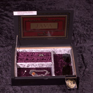 Op de afbeelding: Een zwarte houten kist met een bordeauxrode fluwelen binnenkant, met daarin een ring, zonnebril, armband en speld. Het deksel van de kist is voorzien van de tekst "JAVA" en "Drew Estate". Een zilveren ketting is aan de kist bevestigd.