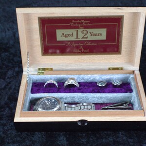 May include: A wooden box with a hinged lid, open to reveal a watch, two rings, two cufflinks, and a tie clip. The box is labeled "Aged 12 Years - A Signature Collection by Riskey Patel".