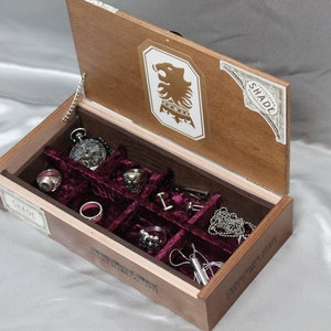 May include: A wooden box with a red velvet interior and compartments for storing jewelry. The box is open and the lid is propped up. The lid has a label that says "SHADE" and a lion crest.