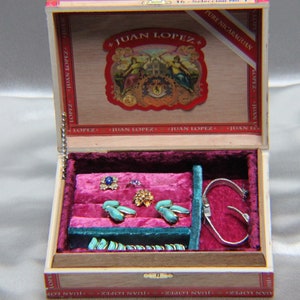 May include: An open wooden box with a red velvet interior. The box is lined with compartments and contains various jewelry pieces, including rings, earrings, and a bracelet. The box is labeled "JUAN LOPEZ" on the top.